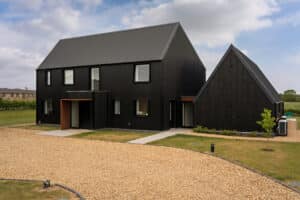 Modern two-story house with black exterior and pitched roofs, gravel driveway, manicured lawn, and cloudy sky in the background.