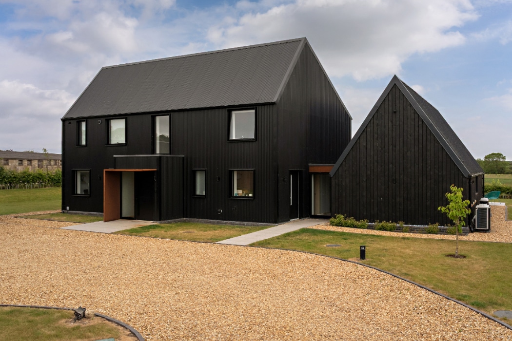 Modern two-story house with black exterior and pitched roofs, gravel driveway, manicured lawn, and cloudy sky in the background.