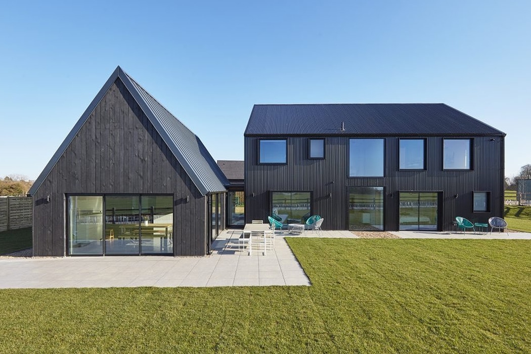 Modern two-story house with black exterior, large windows, and slanted roofs; lawn and patio with outdoor furniture in the foreground.