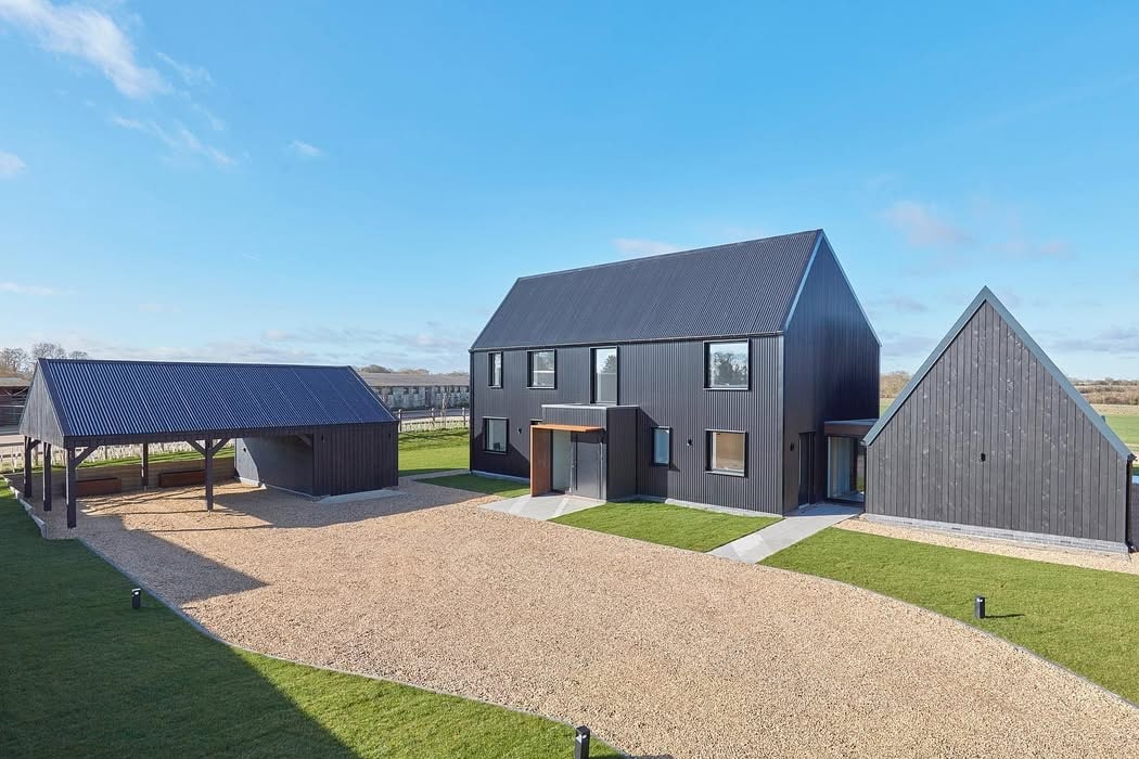 A modern black house with large windows, a separate garage or barn, and a triangular annex, all surrounded by gravel paths and green lawns under a clear blue sky.