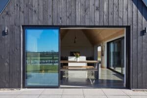 A modern house with dark wood siding and a large sliding glass door opening to a dining table with flowers, overlooking an outdoor field.