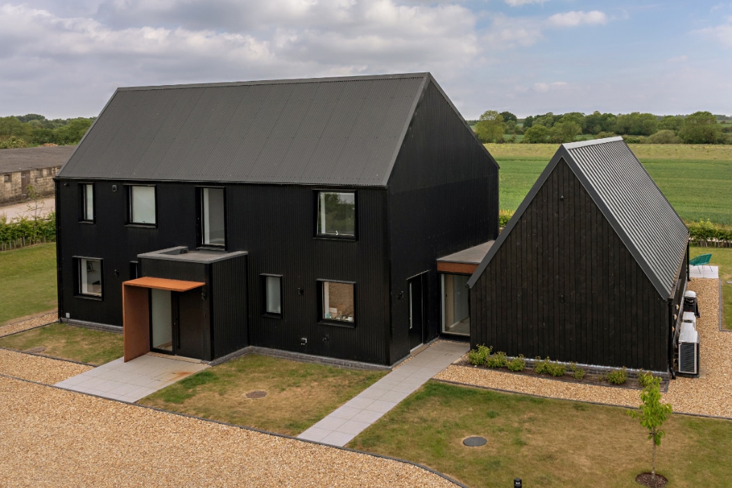 A modern black house with a pitched roof and large windows, connected to a smaller building, set in a rural landscape with fields and cloudy sky.