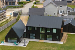 Aerial view of a modern black house with large windows and an adjacent outbuilding, set on a green lawn with patio furniture and surrounded by other homes.