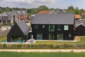 A modern black house with large windows and an adjoining building, set in a residential area with other homes and greenery visible in the background.