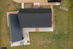Aerial view of a modern house with dark roof, surrounding patio, outdoor furniture, and green lawn. Gravel paths border parts of the property.