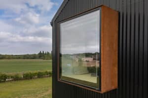 Large, box-shaped window with a rust-colored frame protruding from a black corrugated metal wall, overlooking a grassy field and trees under a partly cloudy sky.
