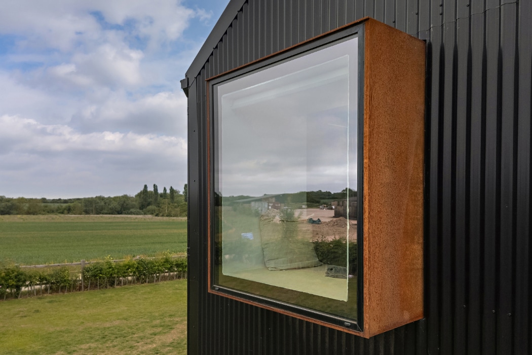 Large, box-shaped window with a rust-colored frame protruding from a black corrugated metal wall, overlooking a grassy field and trees under a partly cloudy sky.