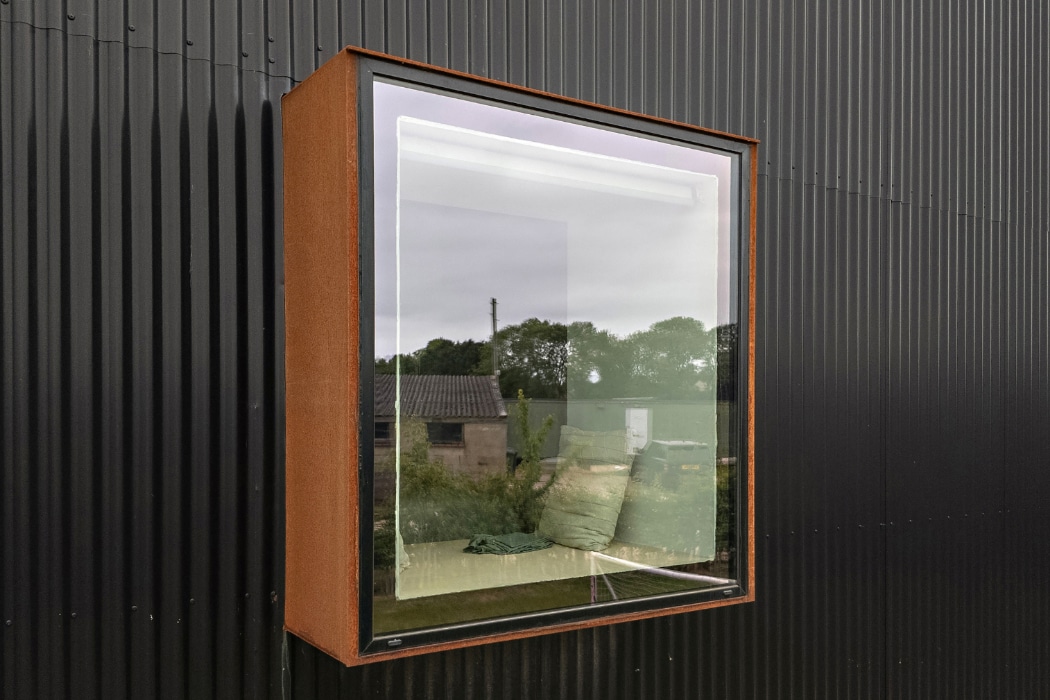 A box-shaped window with a rusty metal frame extends from a black corrugated metal wall, reflecting an outdoor scene with trees, buildings, and cloudy sky.