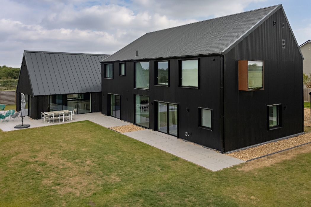 Modern black two-story house with large windows and an adjoining single-story section, situated on a lawn with a stone patio and outdoor dining area.