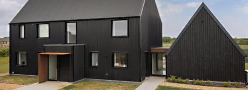 A modern black house with large windows, a sloped roof, and an attached angular extension, set in a grassy landscape under a partly cloudy sky.