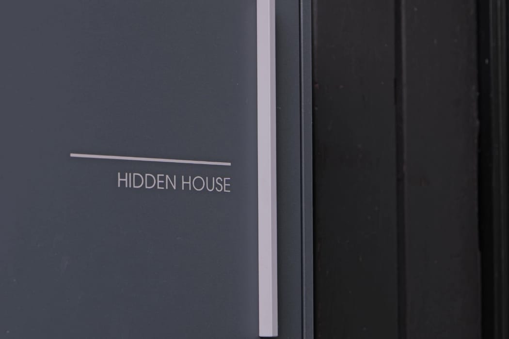 A close-up of a dark gray sign with the words "HIDDEN HOUSE" in white capital letters, mounted next to a black surface.