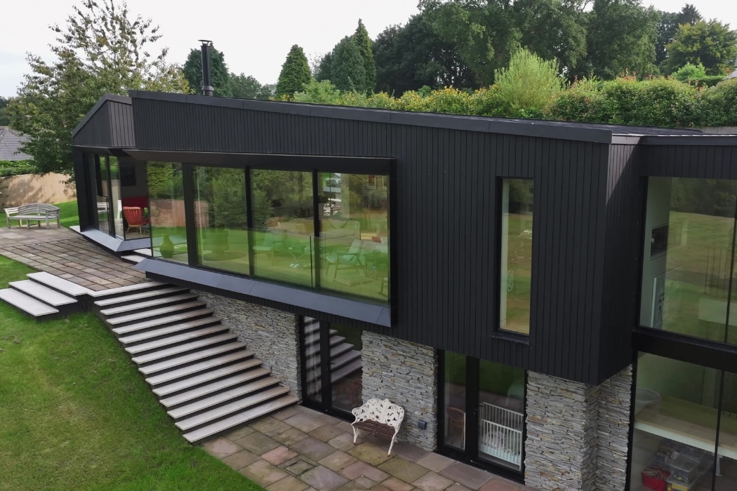 Modern two-story house with large glass windows, stone and dark panel exterior, and outdoor stairs surrounded by greenery.