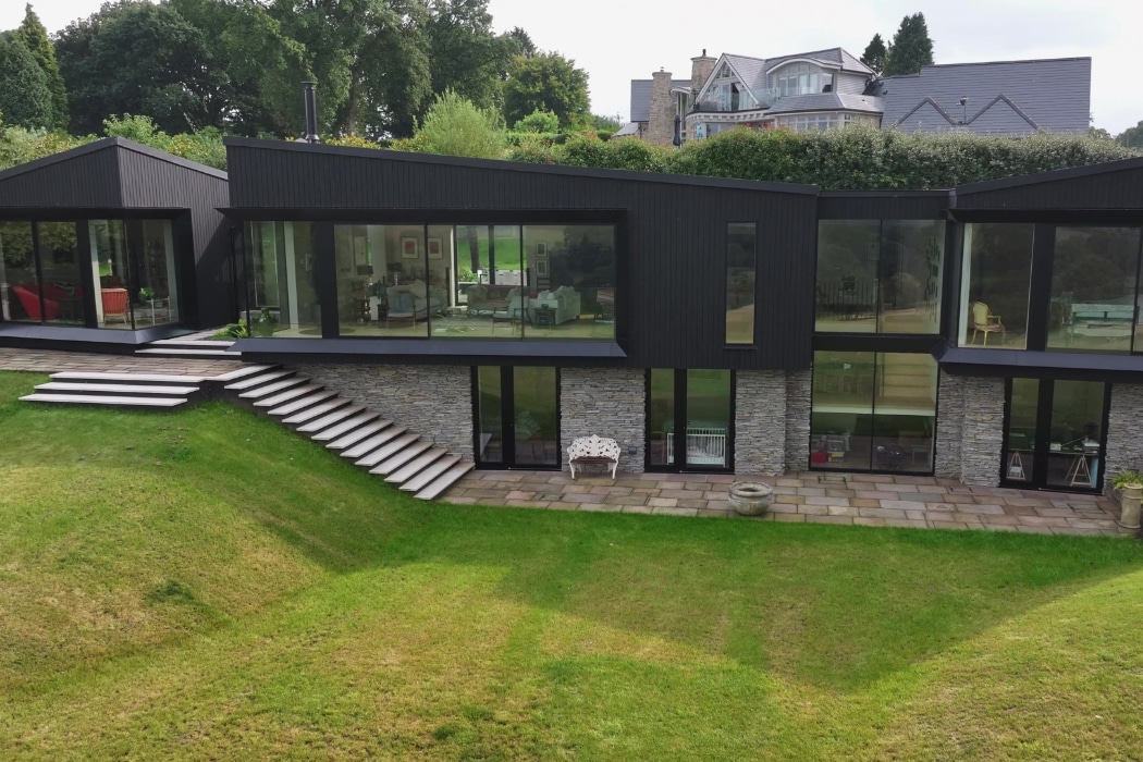 Modern two-story house with dark exterior, large glass windows, stone lower level, wide steps, and a landscaped grassy yard.
