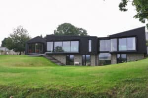 Modern house with large floor-to-ceiling windows, dark siding, and stone accents, situated on a grassy hill with trees in the background.