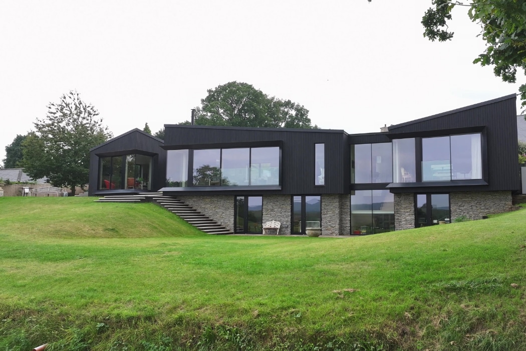 Modern house with large floor-to-ceiling windows, dark siding, and stone accents, situated on a grassy hill with trees in the background.