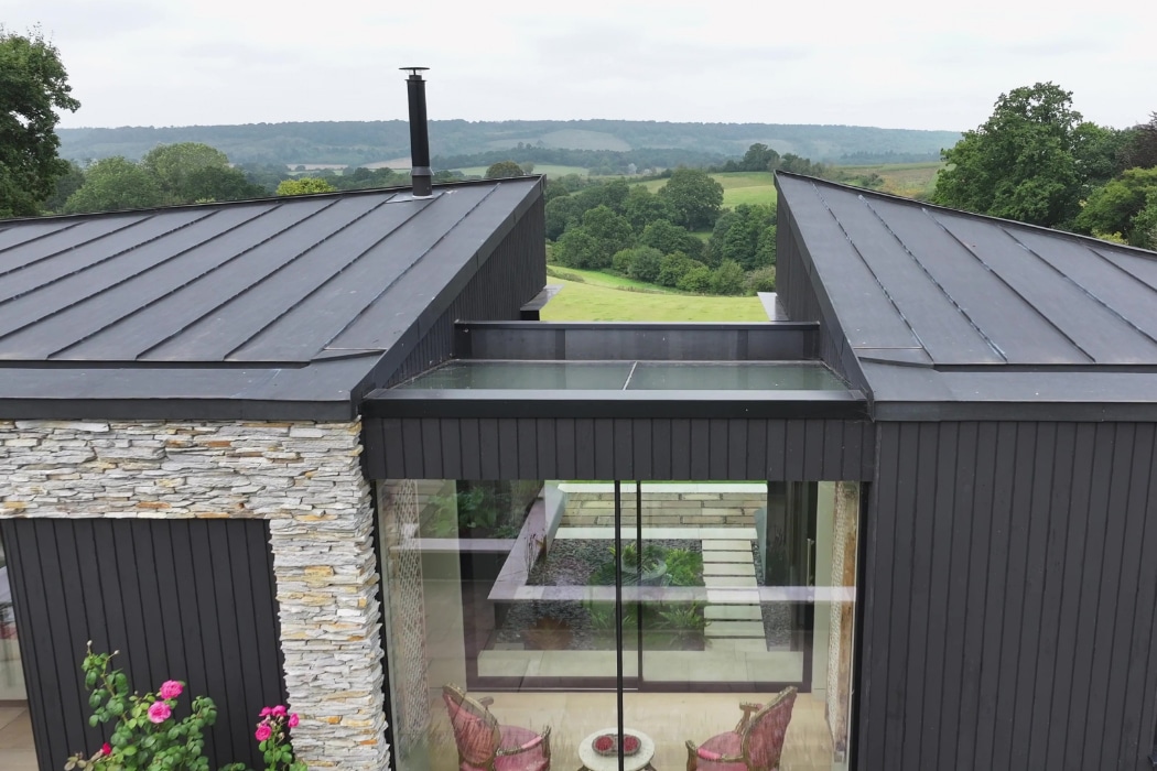 Modern house with dark vertical siding and stone accents, flat metal roof, and large glass windows overlooking a green rural landscape.