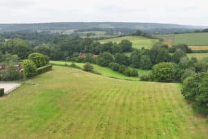 A wide grassy field with scattered trees, bordered by hedges and rolling hills in the background under a cloudy sky.