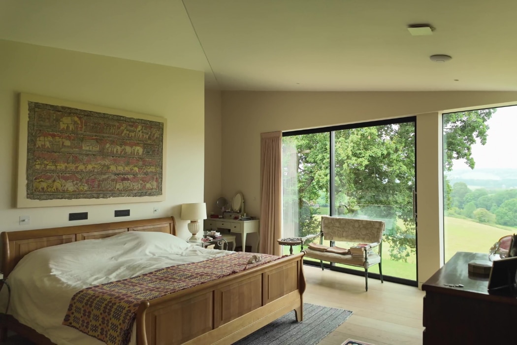 Bedroom with a wooden bed, wall art, a side table with a lamp, a bench, and a large glass window overlooking trees and a grassy landscape.