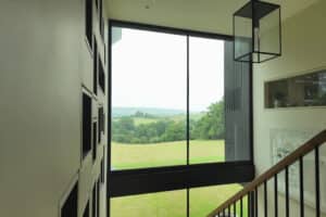 A large window in a modern building overlooks a green landscape with trees and rolling hills; stairs and a geometric light fixture are visible inside.