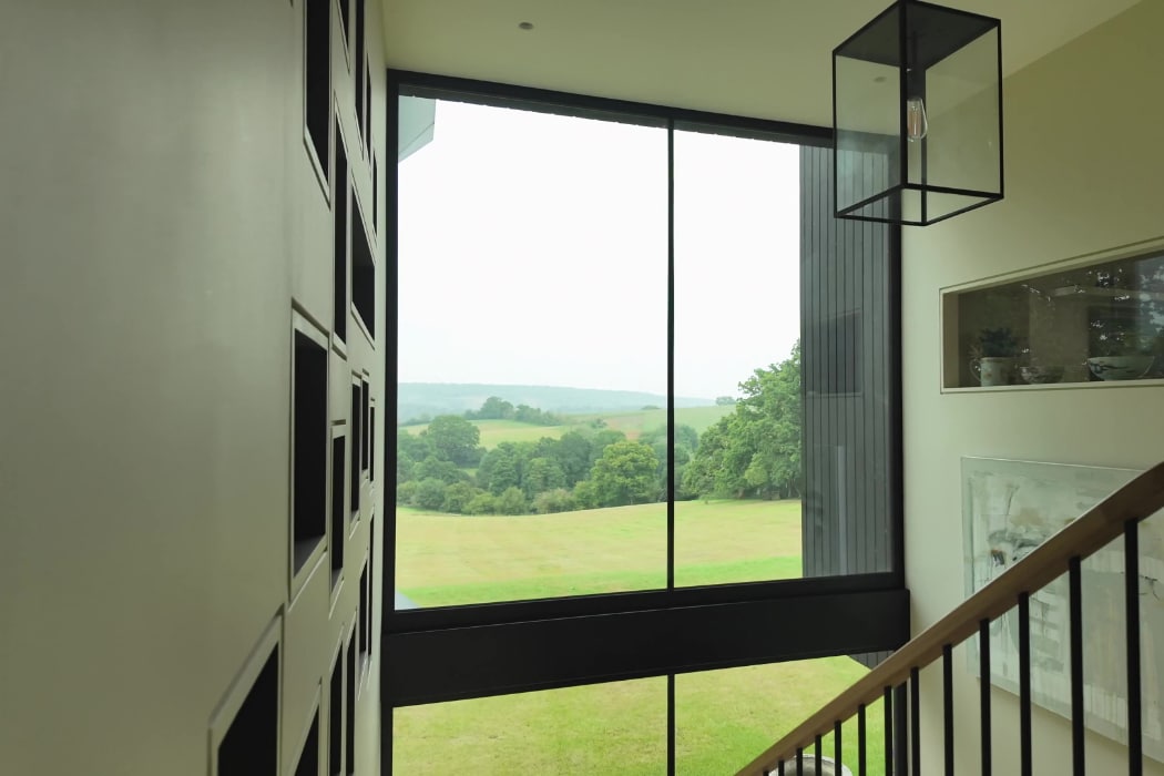 A large window in a modern building overlooks a green landscape with trees and rolling hills; stairs and a geometric light fixture are visible inside.