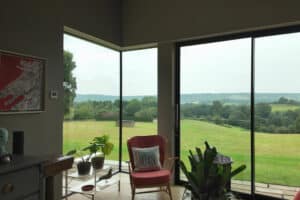 A modern living room with large floor-to-ceiling windows overlooks a green countryside landscape. A pink chair, houseplants, and artwork are visible in the room.