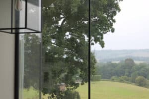 Large window overlooking a green landscape with trees and rolling hills under a cloudy sky; part of a modern light fixture is visible indoors.