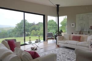Modern living room with large floor-to-ceiling windows overlooking a green landscape, beige sofas, a patterned rug, and contemporary decor.