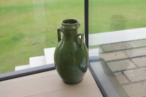 A large green ceramic vase with two handles sits on a light wood floor near a glass window, overlooking a grassy area and stone steps outside.