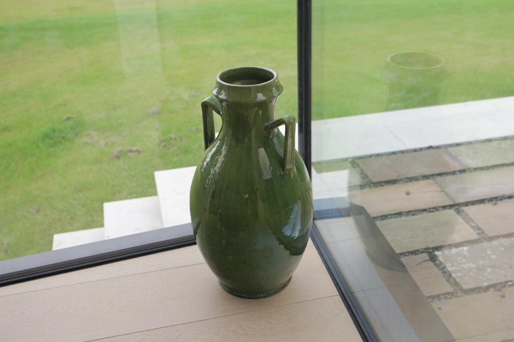 A large green ceramic vase with two handles sits on a light wood floor near a glass window, overlooking a grassy area and stone steps outside.