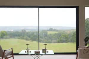 Large window in a modern living room overlooks a green, rolling landscape with trees and distant hills under a cloudy sky.