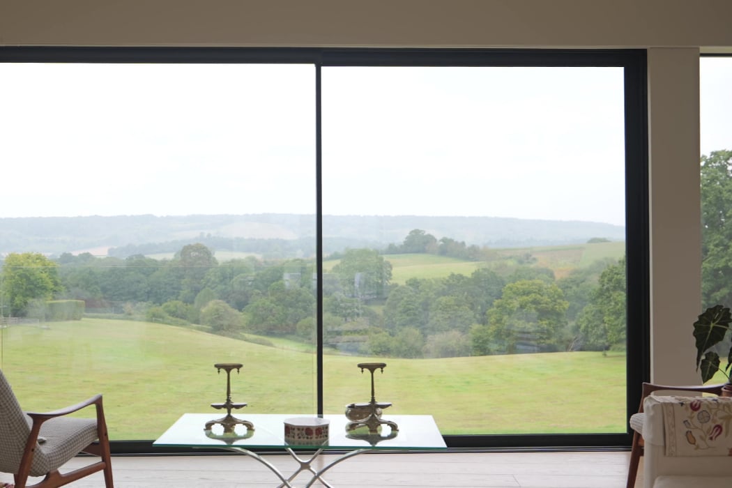 Large window in a modern living room overlooks a green, rolling landscape with trees and distant hills under a cloudy sky.