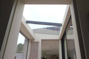 View looking up at a modern building’s glass ceiling with a horizontal beam, large windows, and a lattice panel on the left side.