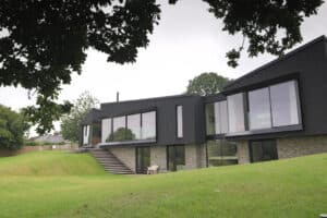 Modern house with large glass windows and dark exterior panels, built on a sloped lawn with stone accents and surrounded by trees and greenery.