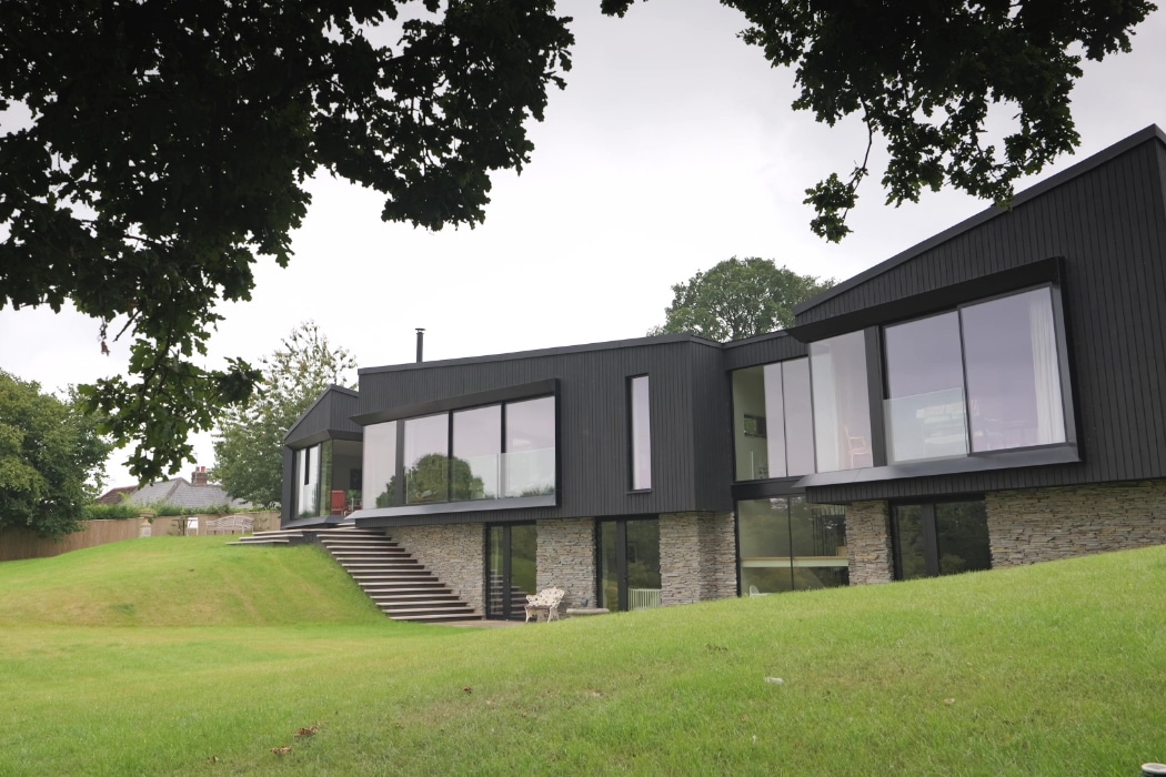 Modern house with large glass windows and dark exterior panels, built on a sloped lawn with stone accents and surrounded by trees and greenery.