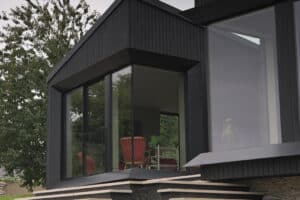 Close-up of a modern house with large floor-to-ceiling windows, black exterior paneling, and a red chair visible inside near a tree.