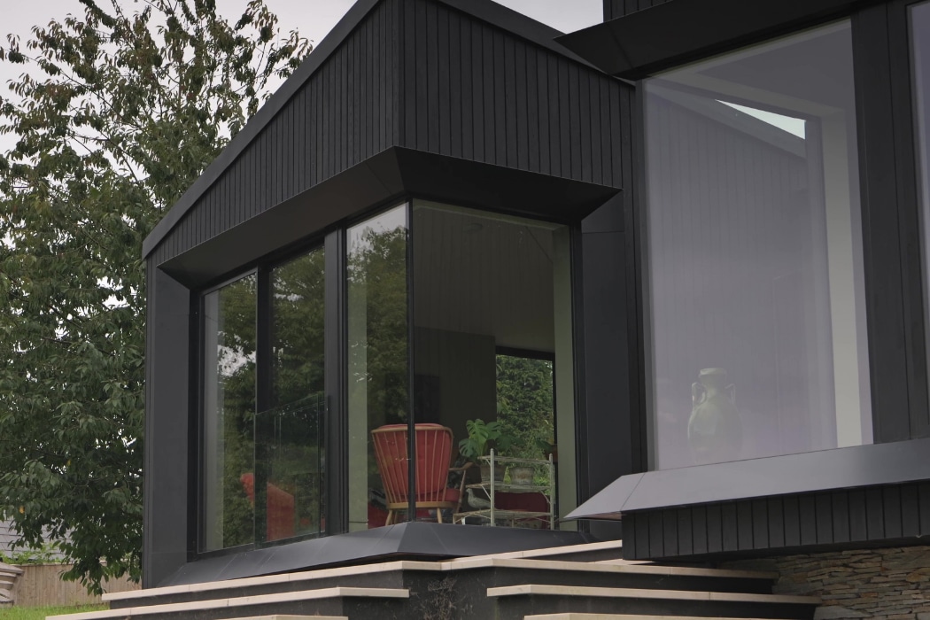 Close-up of a modern house with large floor-to-ceiling windows, black exterior paneling, and a red chair visible inside near a tree.