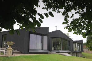 Modern black house with large glass windows and angular rooflines, surrounded by grass and partially framed by tree branches.