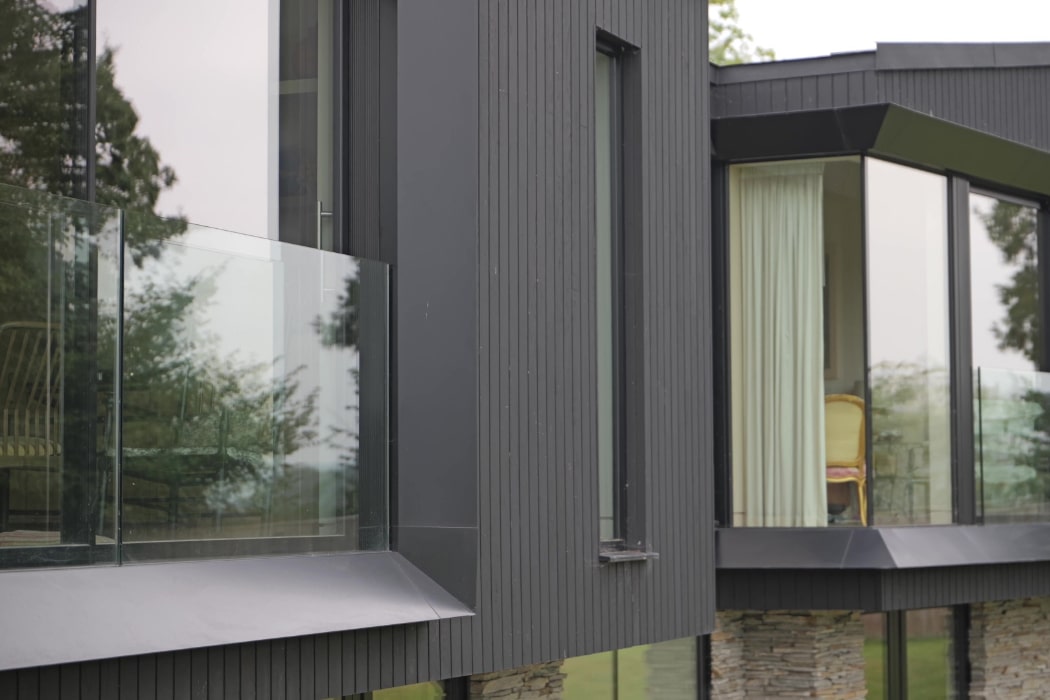 Modern house exterior with dark vertical siding, large glass windows, and stone supports, showing reflections of trees and partial view of indoor furniture.