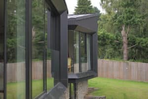 Modern house with large glass windows, stone base, and angular design, overlooking a grassy yard with trees and a wooden fence in the background.