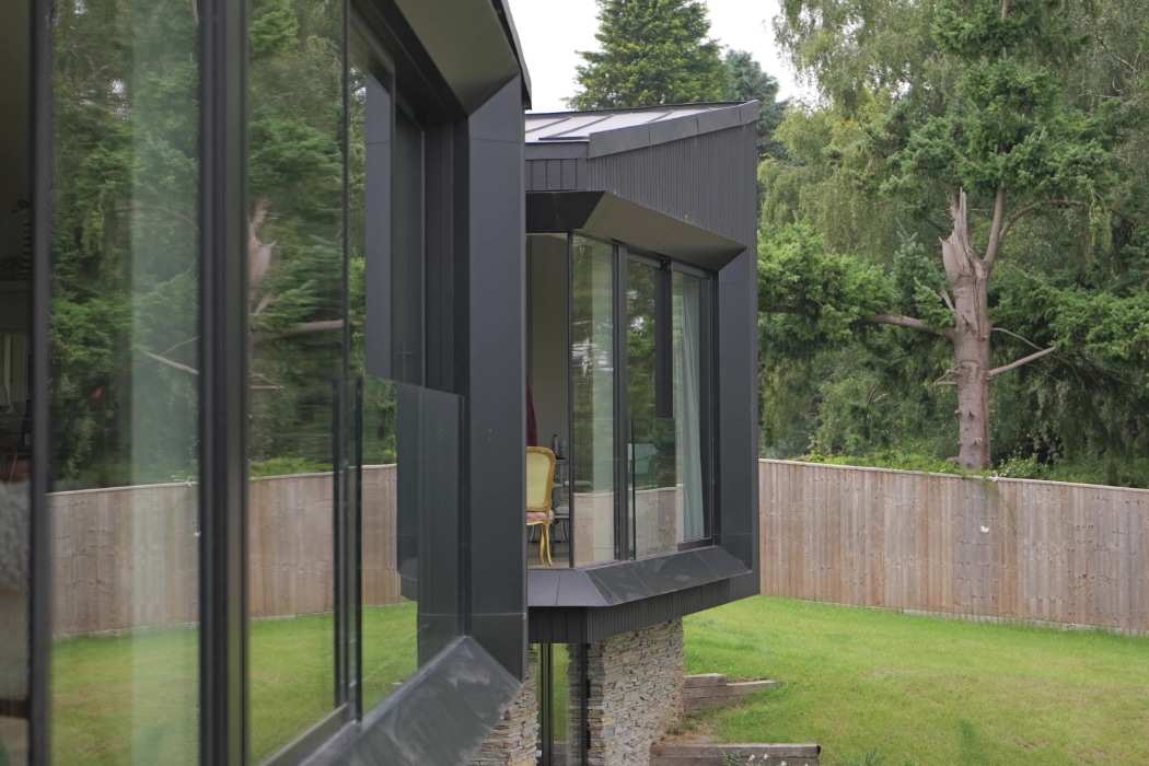Modern house with large glass windows, stone base, and angular design, overlooking a grassy yard with trees and a wooden fence in the background.