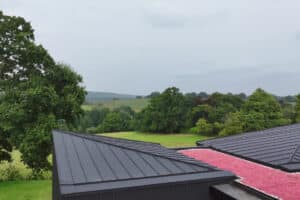 A modern building with a black metal roof and a section covered in pink foliage, set against a lush green landscape and distant hills.