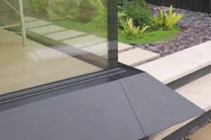 Close-up of a corner glass wall next to a paved stone walkway and landscaped garden with green plants and dark mulch.