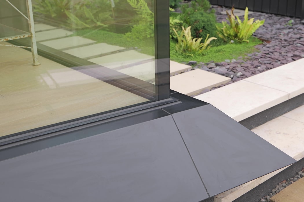 Close-up of a corner glass wall next to a paved stone walkway and landscaped garden with green plants and dark mulch.