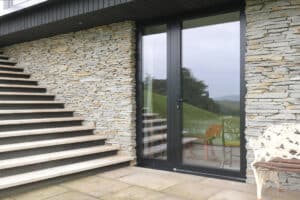 Glass patio door with black frame set in a stone wall, adjacent to an outdoor staircase and a white metal bench on a tiled patio.