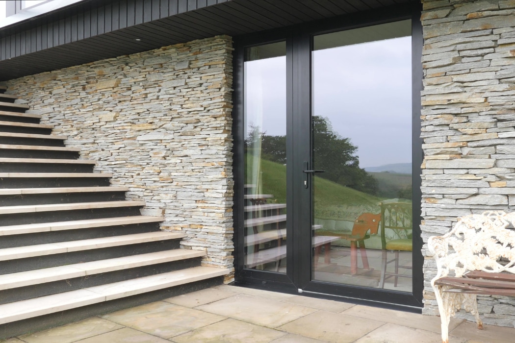 Glass patio door with black frame set in a stone wall, adjacent to an outdoor staircase and a white metal bench on a tiled patio.