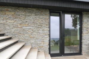 Stone wall with beige steps leading to a glass door framed in black; the door reflects outdoor scenery and shows indoor chairs.