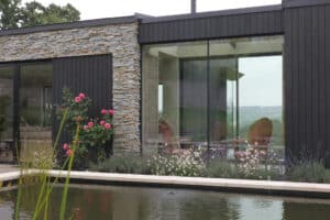 Modern house with stone and dark wood exterior, large glass windows, reflecting garden with blooming flowers and a pond in the foreground.
