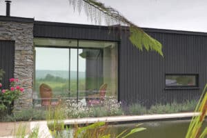 Modern house with dark wood siding, large glass window, and stone feature wall, overlooking a pool and garden with flowers; hills visible in the background.