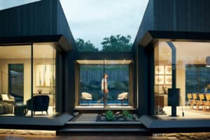 Modern home exterior at dusk with large glass windows showing indoor living and dining areas; a person walks through the central corridor between the two sections.