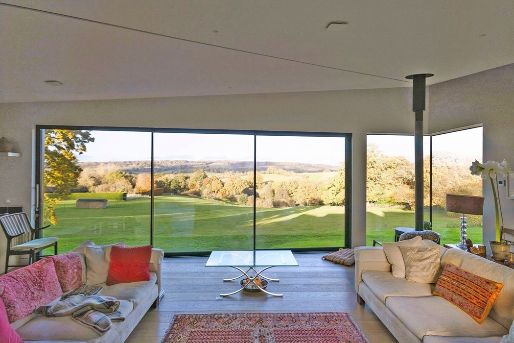 Modern living room with large floor-to-ceiling windows offering a panoramic view of a green lawn and trees under a clear sky.